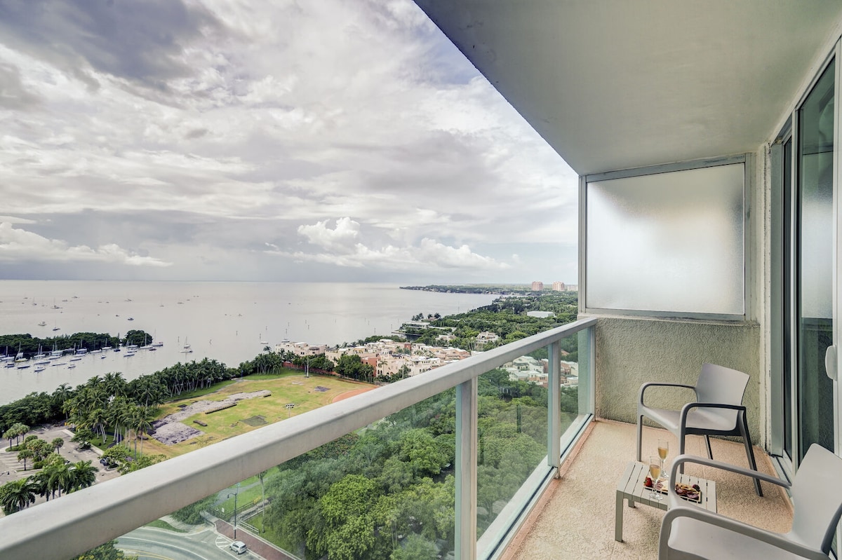 A balcony view showcases a bay landscape, featuring Biscayne Bay and sailboats. Two outdoor chairs are positioned for relaxation, overlooking greenery and the water. The sky is filled with clouds, suggesting a serene ambiance.