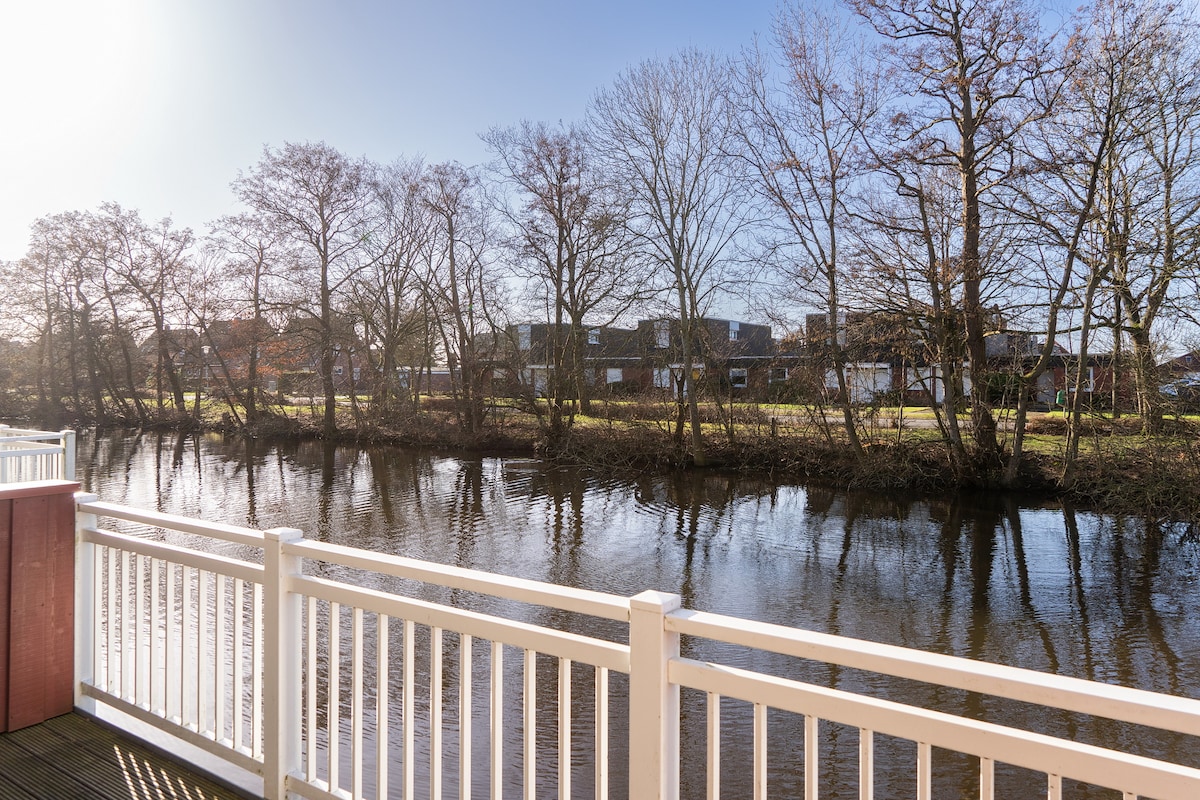 A view of a tranquil waterway is presented, bordered by bare trees reflecting softly on the surface. The scene captures the natural beauty of the surroundings, framed by a wooden railing that enhances the serene atmosphere.