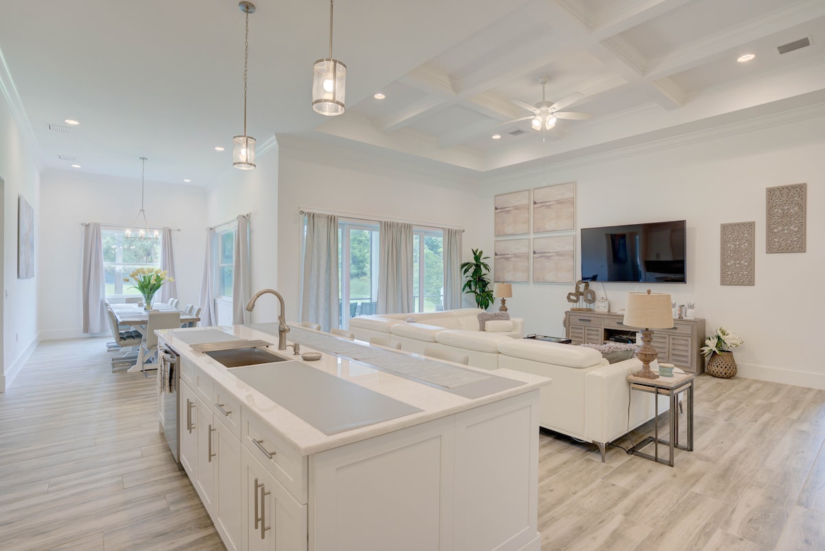 A spacious living area features a coffered ceiling and large windows that let in natural light. An elegant kitchen island with a sink separates the kitchen from the open living space, which includes a white sectional sofa and a wall-mounted television.