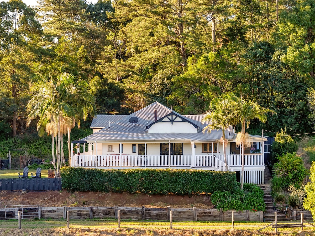 A charming holiday home is nestled among lush greenery, featuring a spacious verandah with railings. Palm trees line the front, and stairs lead to the entrance. The home’s exterior showcases a combination of white walls and a grey roof, creating an inviting atmosphere in a serene setting.