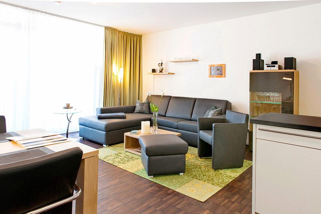 A spacious living area features a sectional sofa with dark upholstery, complemented by an armchair. A light-colored coffee table sits atop a patterned area rug. Soft lighting from the floor lamps illuminates the room, which includes a modern entertainment unit and large windows covered with sheer curtains.