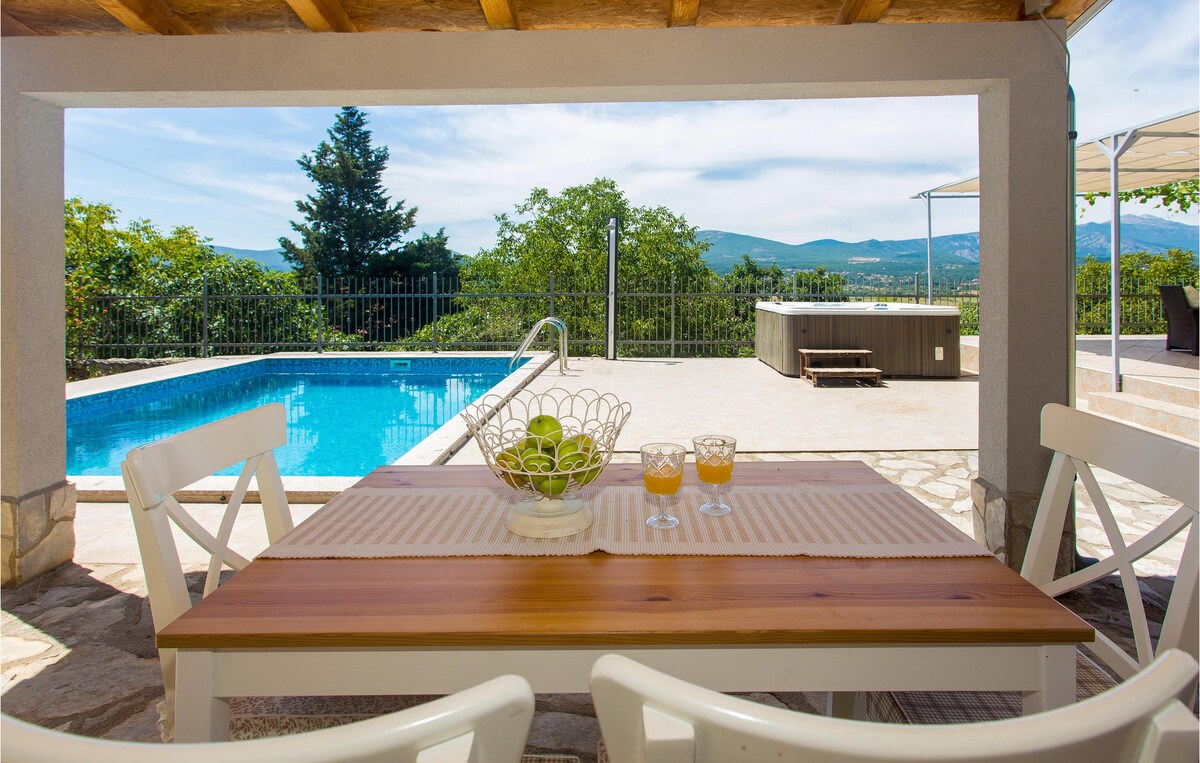 A well-furnished outdoor dining area features a wooden table set with a fruit bowl and two drinks. The private pool is visible in the background, surrounded by a serene landscape of greenery and distant hills.