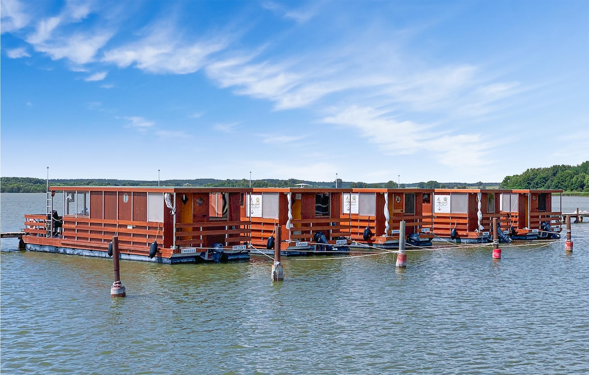 A series of floating cabins are seen anchored on the calm water, surrounded by lush greenery in the distance. The structures feature wooden exteriors and large windows, providing an appealing blend of comfort and nature.