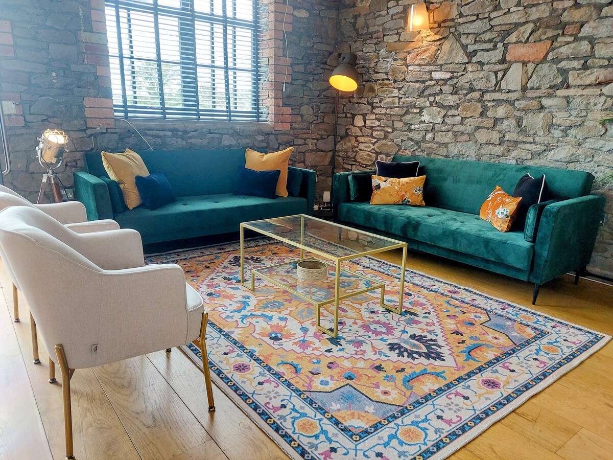 The living area features green upholstered sofas adorned with decorative cushions. A glass coffee table rests atop a colorful area rug, which adds warmth to the wooden floor. Natural light filters through the window, highlighting the textured stone walls.