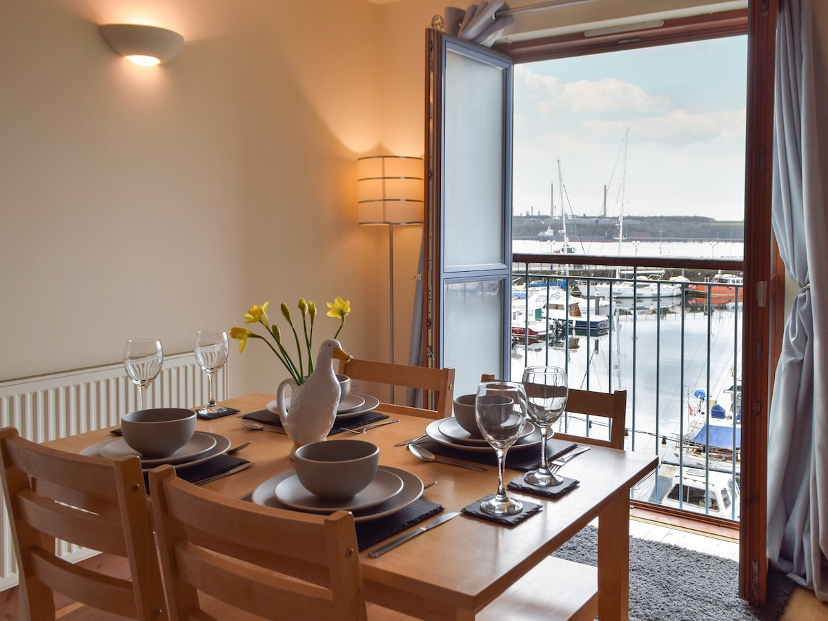 A dining area is presented with a wooden table, set with plates and glasses. Natural light filters through the French doors, leading to a Juliet balcony overlooking the marina. A vase of yellow flowers adds a touch of color to the setting.