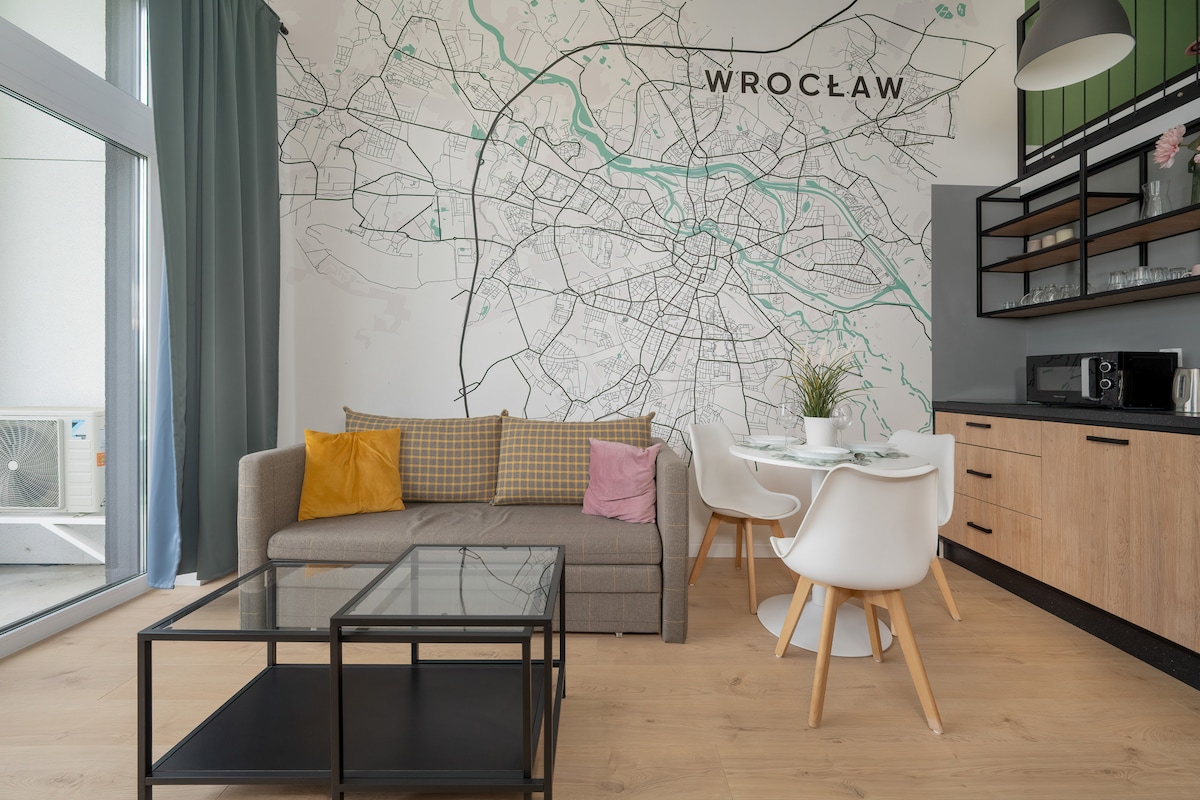A modern living area features a gray sofa with colorful cushions accompanied by a glass coffee table. A dining space with white chairs surrounds a wooden table. A detailed map of Wrocław adorns the wall, enhancing the contemporary design of the room.