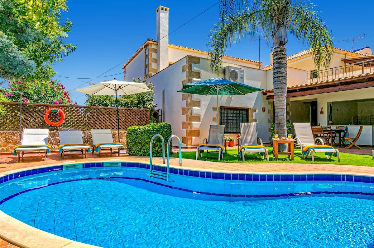 A private swimming pool is featured in the foreground, bordered by lounge chairs and shaded by umbrellas. Lush greenery surrounds the area, with the villa's exterior visible in the background. A seating arrangement is located nearby, providing a relaxing outdoor space.