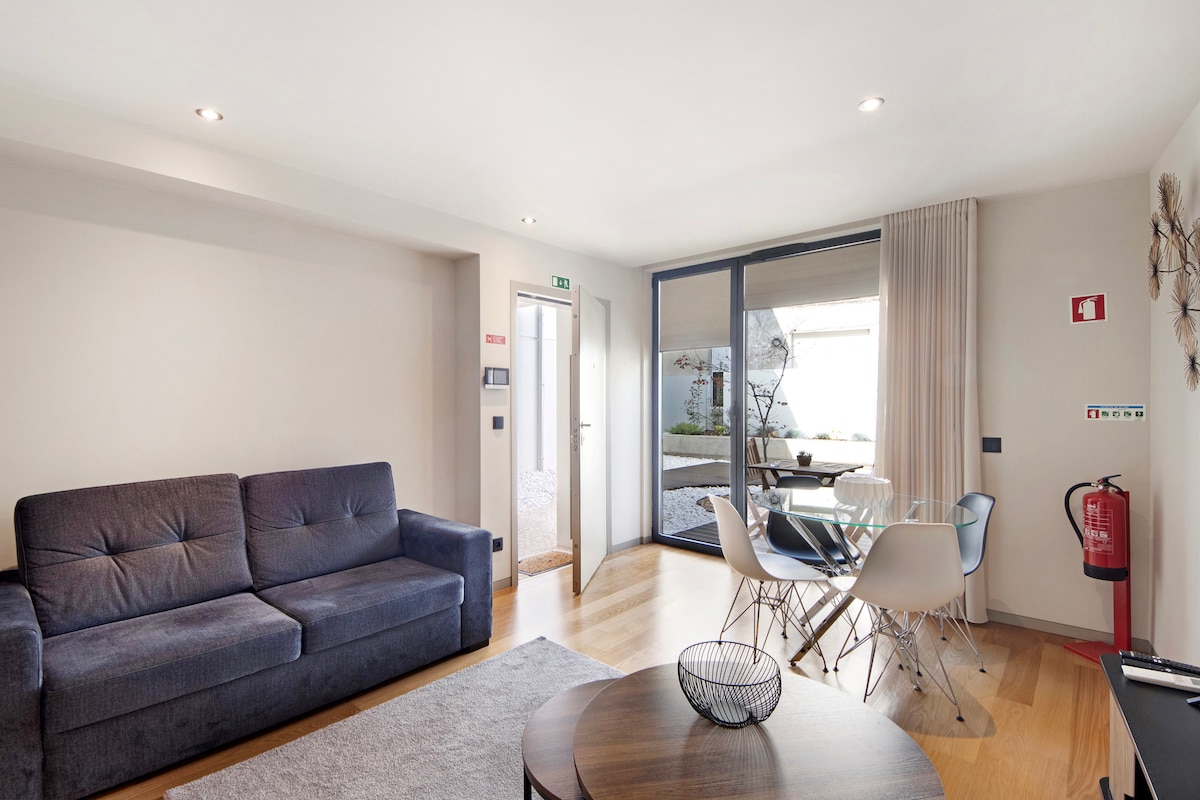 A bright living area features a comfortable sofa, a round dining table surrounded by chairs, and large glass doors leading to an outdoor garden. Natural light fills the space, highlighting the wooden flooring and modern decor.