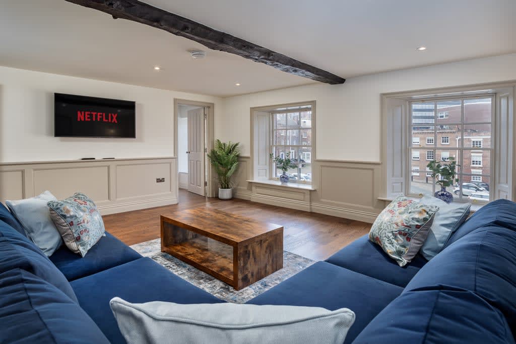 A spacious living room features a large sectional sofa adorned with decorative pillows. A wooden coffee table is centrally positioned, and a wall-mounted Smart TV displays 'NETFLIX.' Two windows allow natural light to brighten the area, complemented by a plant near one window.
