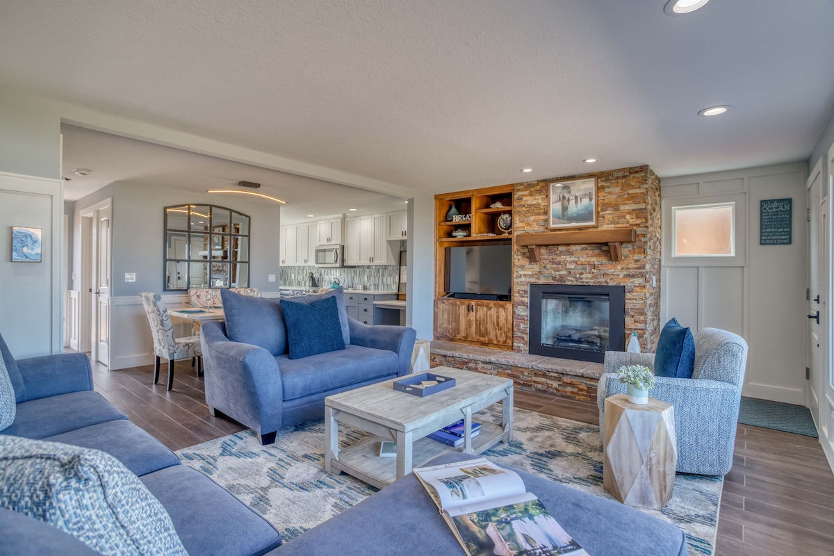 A spacious living room features comfortable blue sofas arranged around a wooden coffee table. A stone fireplace adds warmth, while a mounted TV sits above a rustic wood mantle. Soft lighting enhances the inviting atmosphere, with views of the kitchen and dining area visible in the background.
