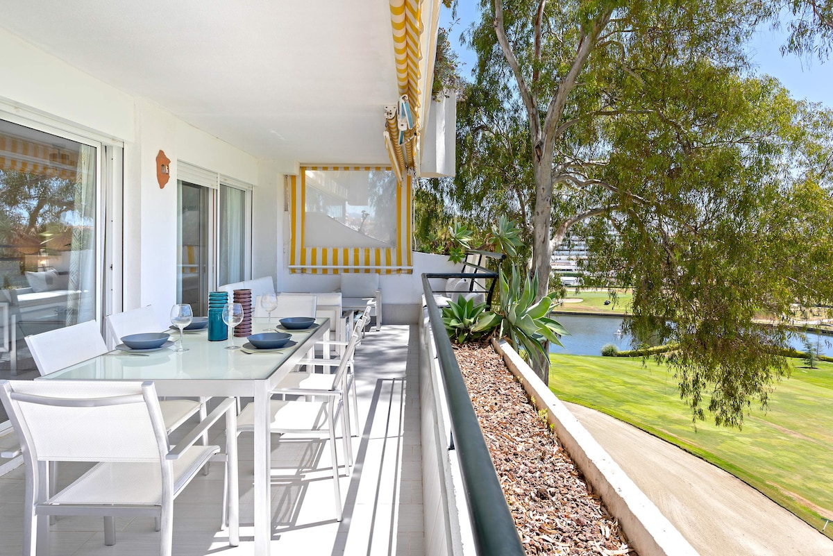 A private terrace is depicted, featuring a dining table set for several guests, surrounded by white chairs. Lush greenery and a golf course view are visible beyond the railing, complemented by a touch of natural light filtering in.