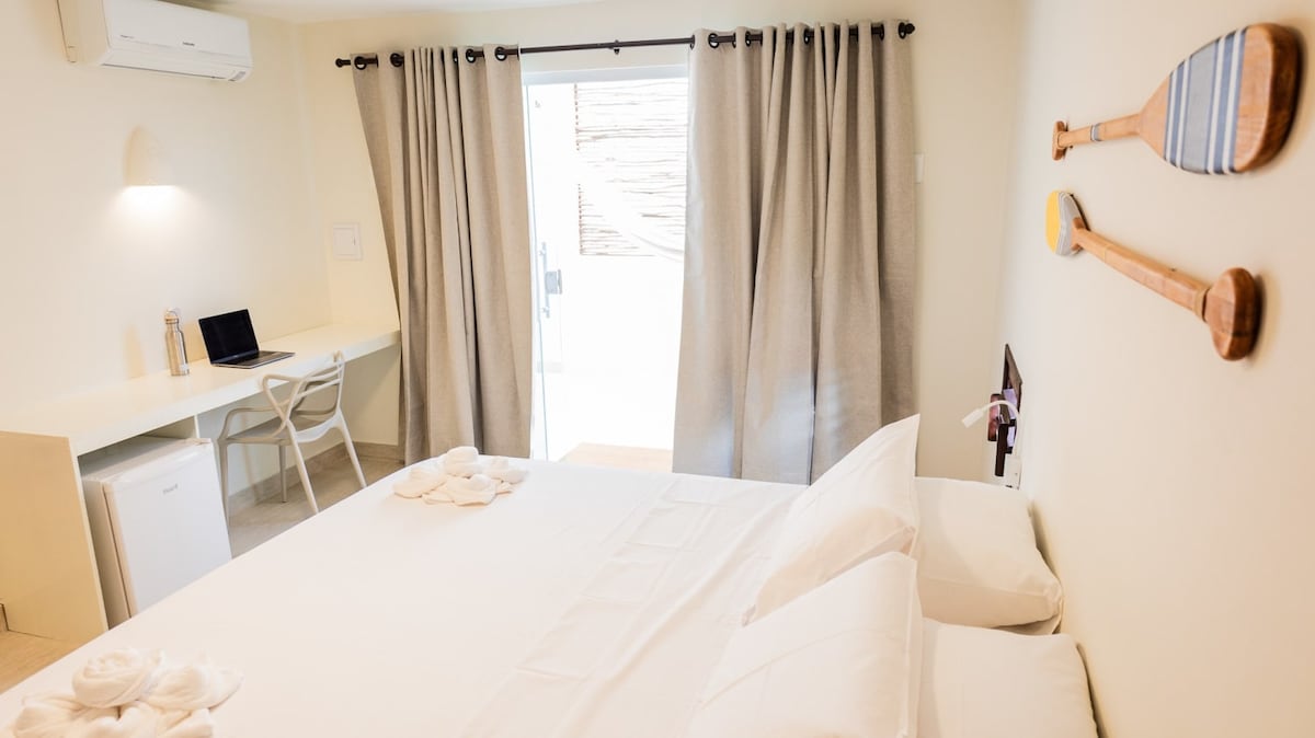 The well-lit room features a spacious bed with neatly arranged linens, a desk, and a mini-fridge. Soft curtains frame a door leading to a private terrace, while wall decor includes wooden paddles, adding a touch of character to the space.