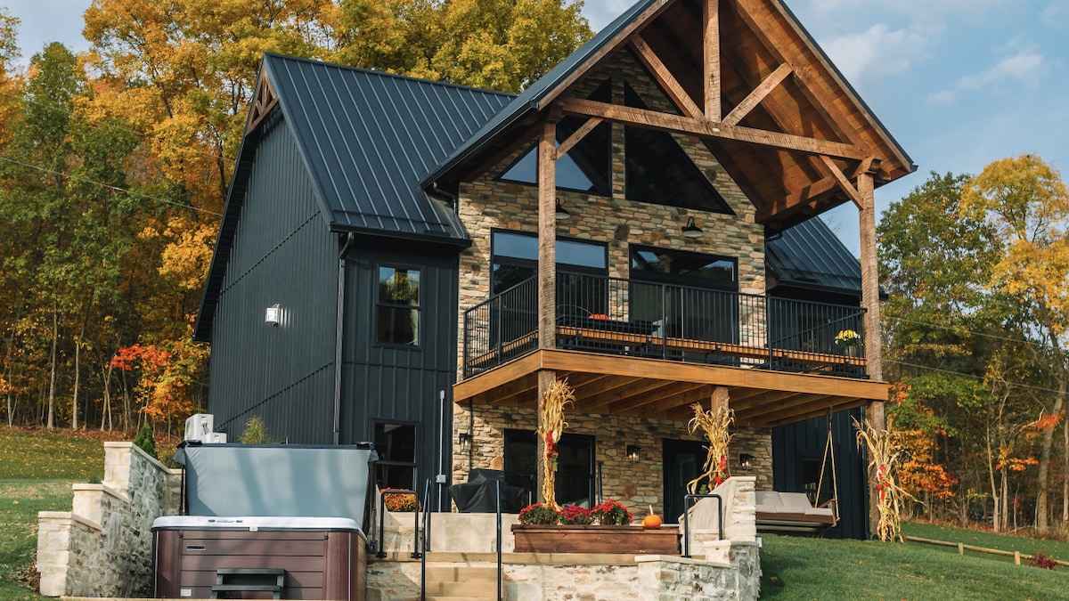 The cabin's exterior showcases a modern design with a combination of stone and sleek black materials. A covered balcony with wooden beams is visible above an inviting hot tub situated on a stone patio, surrounded by lush greenery and autumn foliage.
