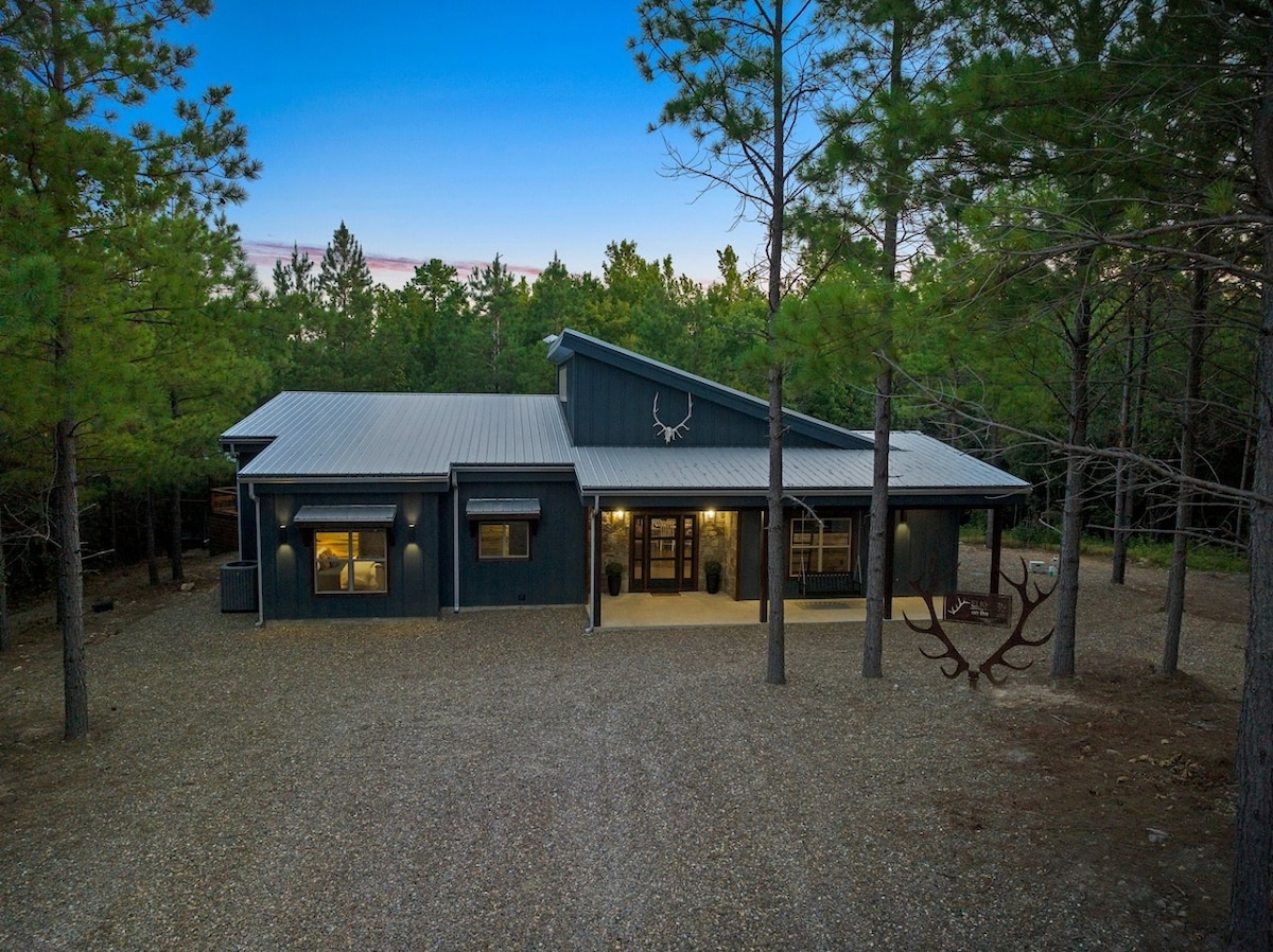 The cabin's exterior is framed by tall pine trees, showcasing a modern design with a metal roof and a welcoming entrance. A gravel driveway leads to the front porch, providing a rustic charm against the serene wooded backdrop.