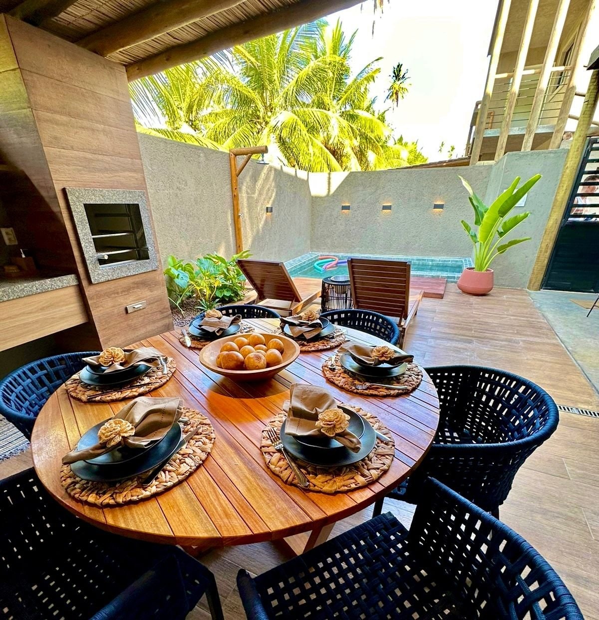 An outdoor dining area features a round wooden table set for six with dark woven chairs. A decorative bowl of fruits sits at the center, surrounded by lush greenery and a glimpse of a private pool in the background.
