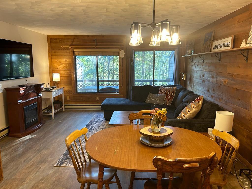 A cozy living space features warm wooden walls and large windows that invite natural light. A sectional sofa offers ample seating, while a round dining table accommodates six. Decorative accents compliment the space, including a TV, a lamp, and a small decorative shelf.