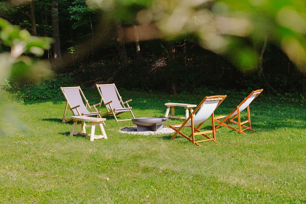 A cozy outdoor seating area features five folding chairs arranged around a circular fire pit, set on a grassy lawn. Surrounded by trees, this tranquil spot invites relaxation and gathering under the open sky.