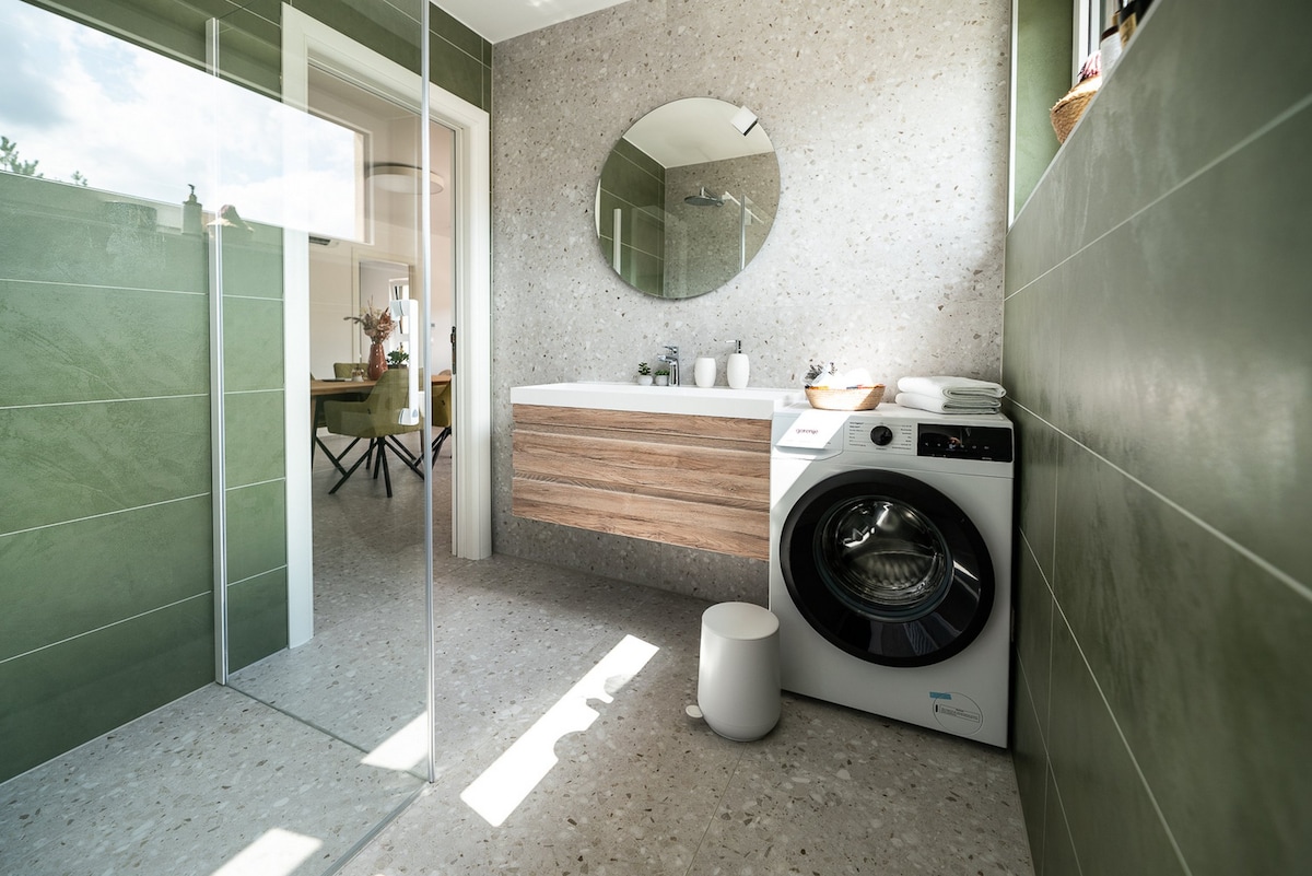 A modern bathroom features a glass shower enclosure and a round mirror above a wooden vanity. A washing machine is positioned next to the vanity, with neatly arranged towels nearby. Natural light filters in, highlighting the textured green walls and elegant tile flooring.