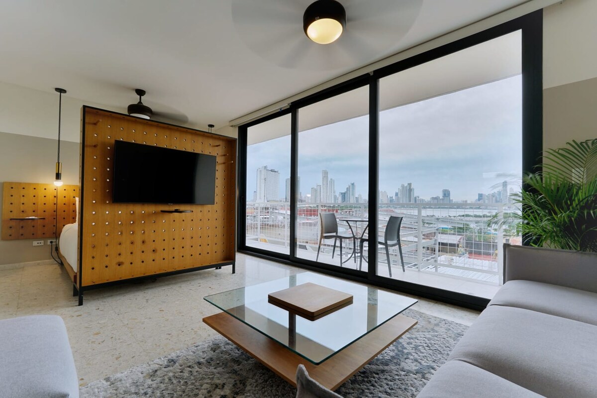 A modern living area is furnished with a comfortable sofa and a glass coffee table. A wooden feature wall supports a wall-mounted television. Floor-to-ceiling windows provide a panoramic view of the city skyline. A small outdoor seating area is visible on the balcony.