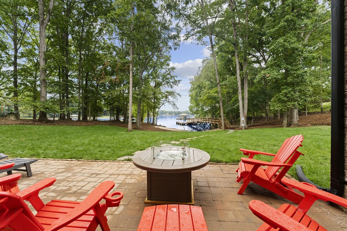 A patio area is shown with vibrant red Adirondack chairs arranged around a fire pit table. Lush green grass frames the space, leading to the lake visible in the background, surrounded by tall trees. A private dock peeks out from the shoreline.