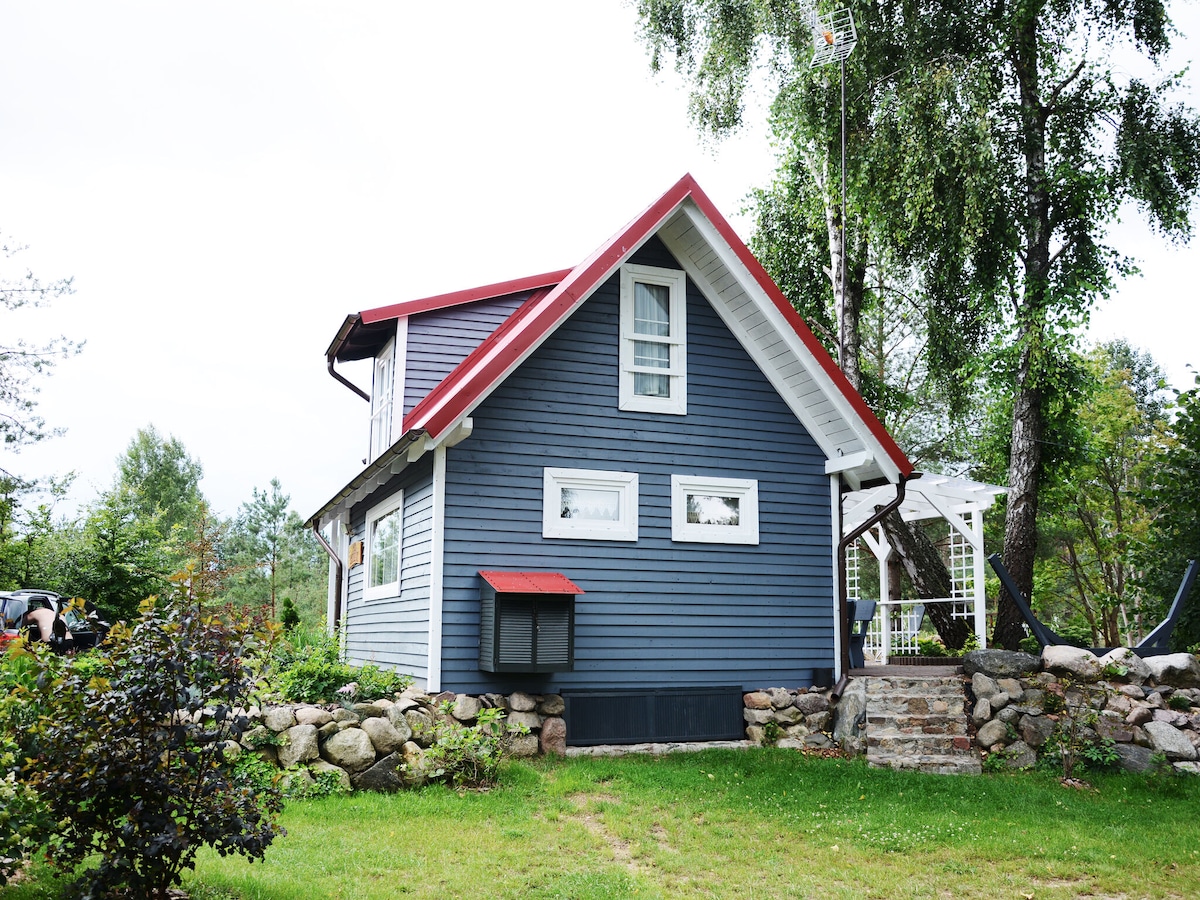 The exterior of a two-story holiday home is visible, featuring a blue facade with white trim and a red roof. A stone wall surrounds the property, and lush greenery is present in the garden, providing a peaceful ambiance.