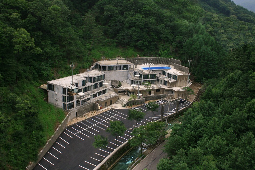 A modern building is nestled among lush greenery, showcasing a unique design with multiple levels. A swimming pool is visible on the terrace, surrounded by a spacious outdoor area. An expansive parking lot is located in the foreground, accommodating numerous vehicles.