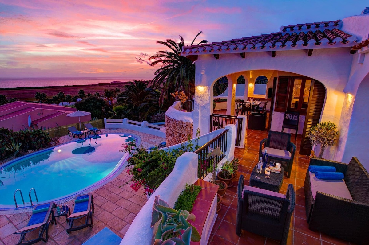 An outdoor seating area is visible next to a private pool, surrounded by lush greenery and palm trees. The sunset creates vibrant colors in the sky, enhancing the tranquil atmosphere. The pool reflects the evening light, and comfortable seating arrangements invite relaxation.