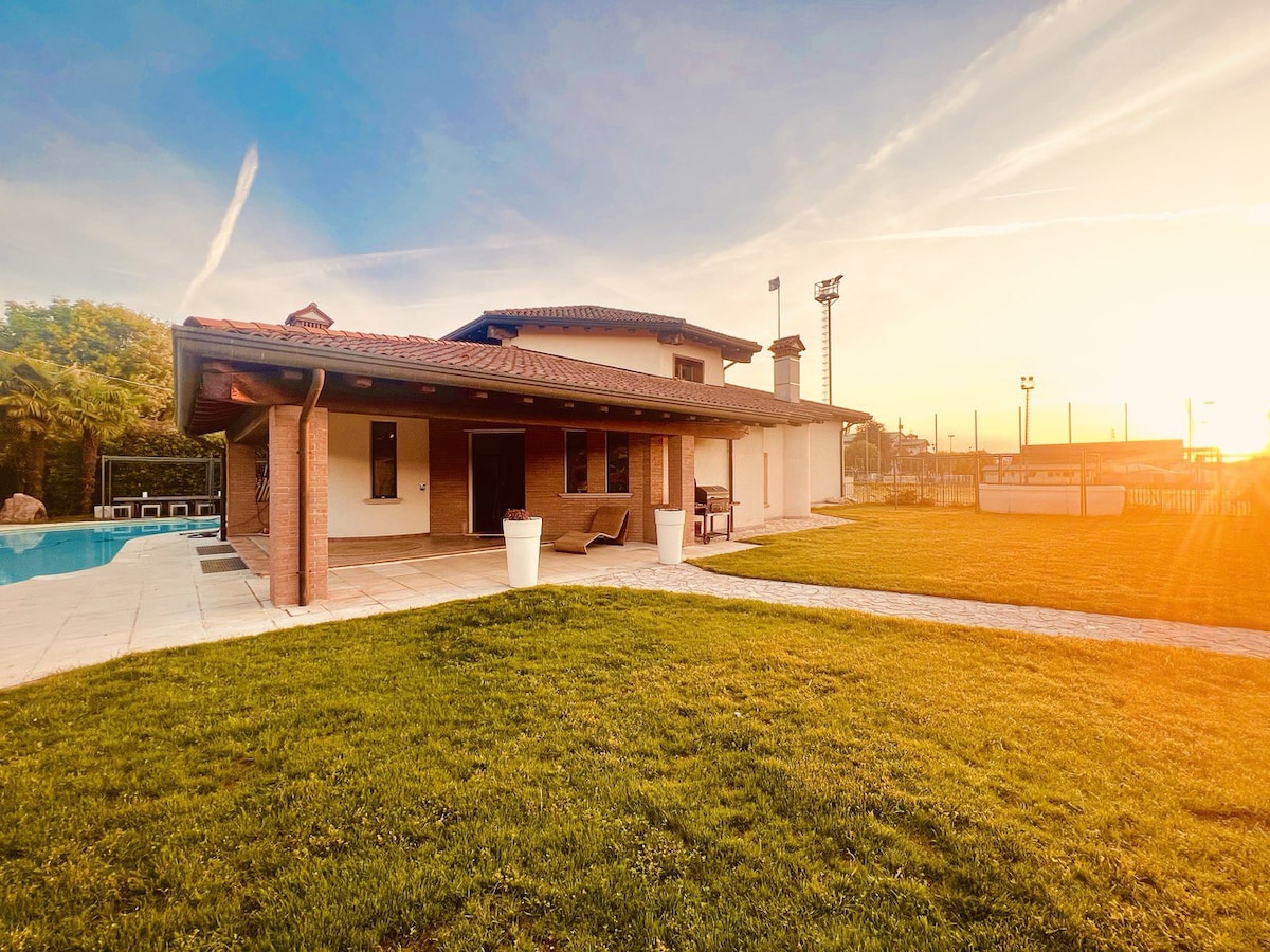 The exterior of a spacious villa is shown, featuring a well-maintained grassy lawn and a patio area. Sunlight bathes the scene, highlighting the house's contemporary architecture and surrounding amenities, including views of sports fields in the background.