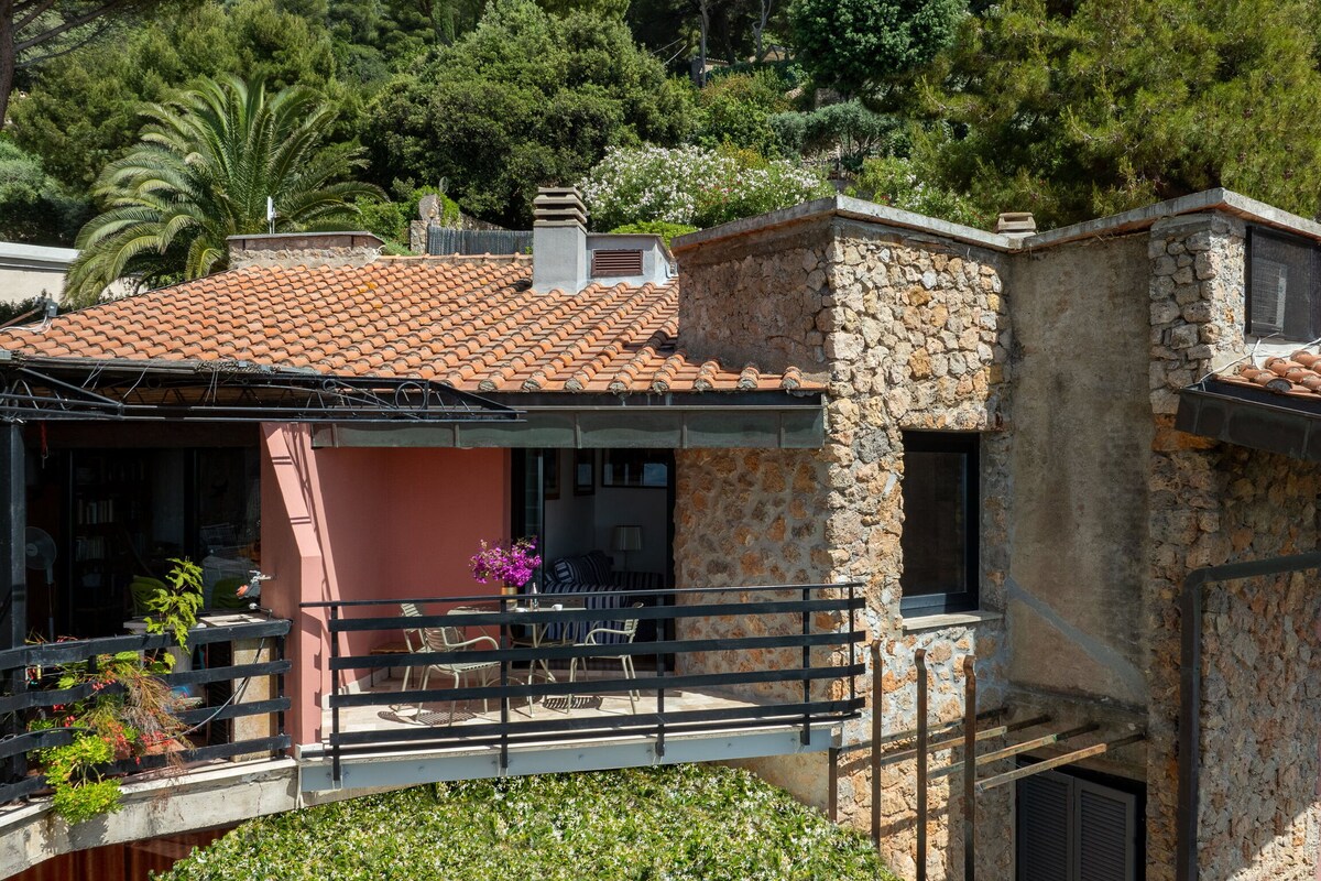 A spacious balcony with a railing is furnished with a small table and chairs, surrounded by lush greenery. The building's exterior features a combination of stone and pink walls, and a red-tiled roof can be seen in the background under clear skies.