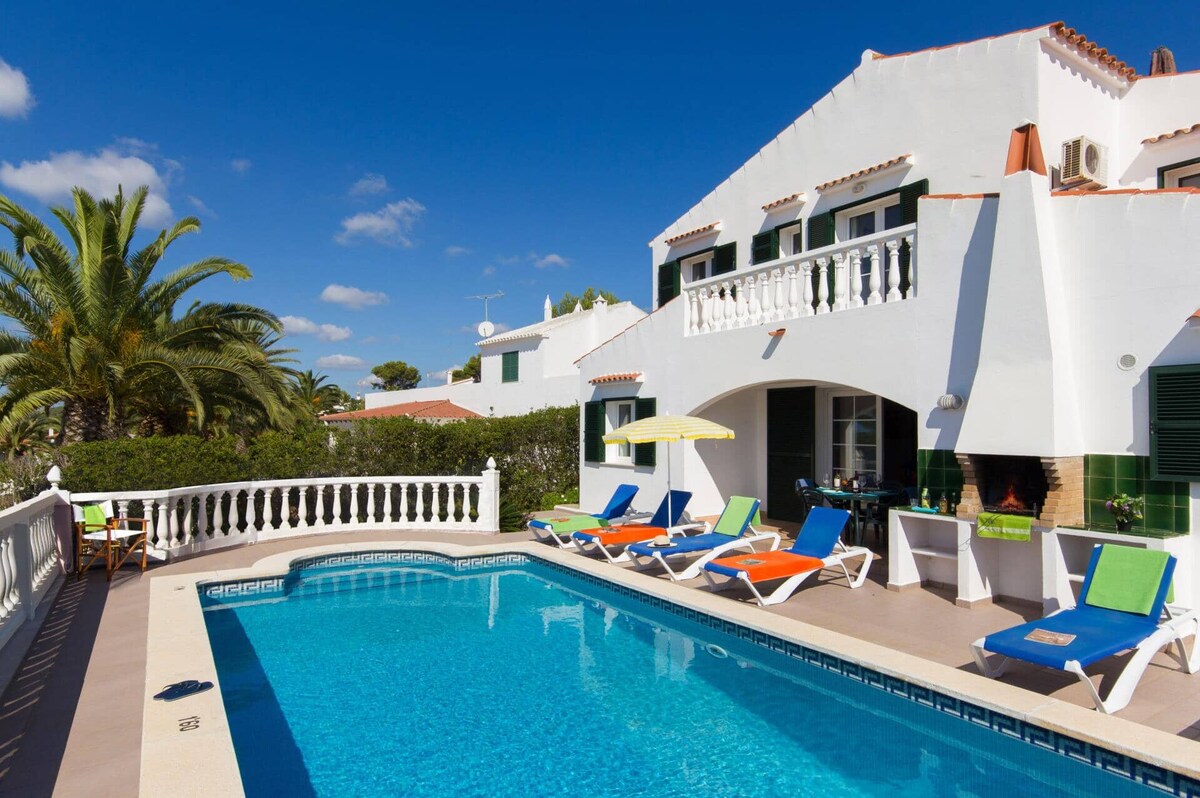 A bright villa exterior is highlighted by a sparkling pool surrounded by colorful lounge chairs. Palm trees provide shade, and a covered seating area is seen in the background. The clear blue sky enhances the inviting atmosphere of the outdoor space.
