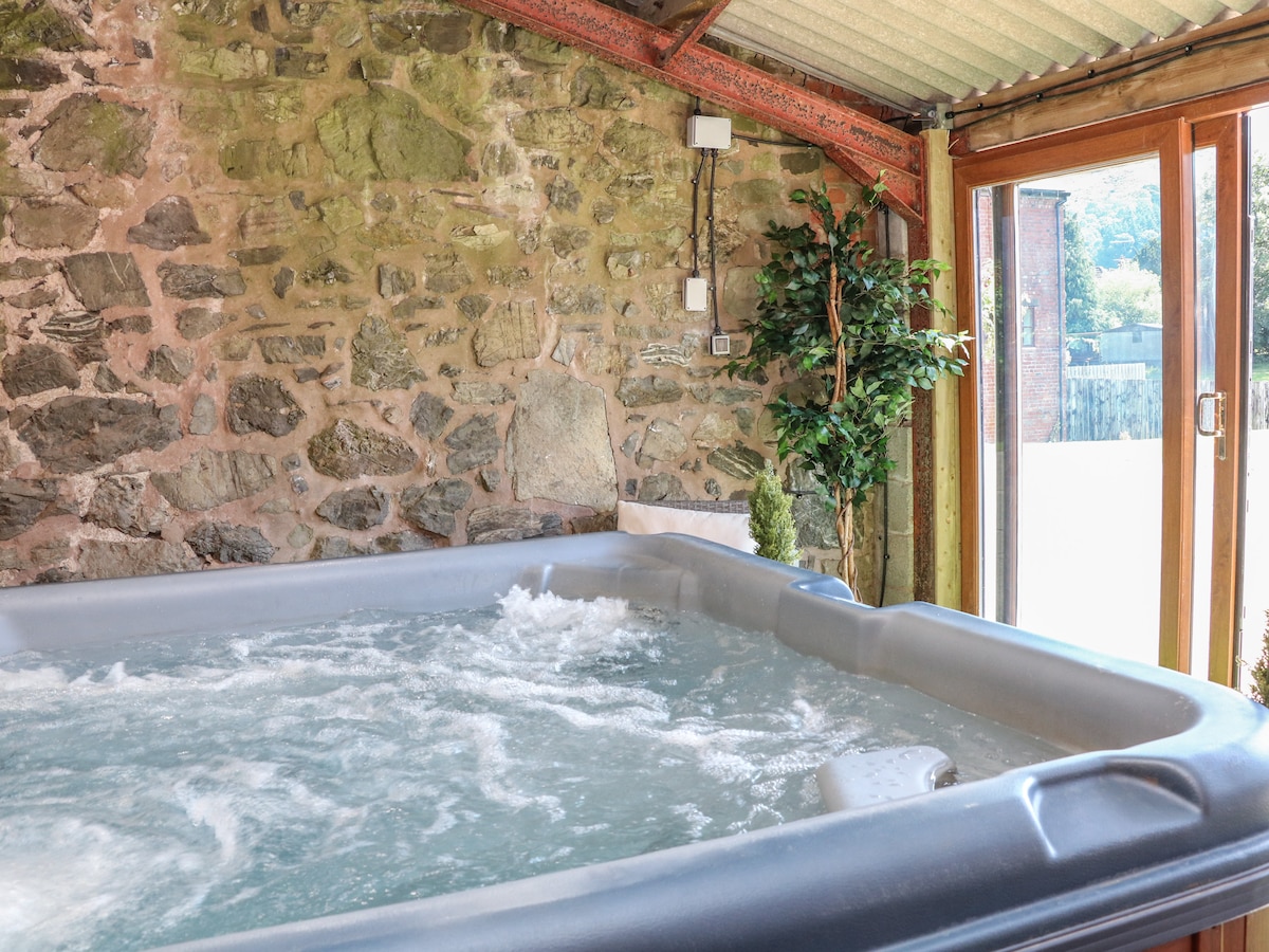 A spacious hot tub sits within a rustic room, featuring stone walls and large windows allowing natural light. Lush greenery is placed nearby, adding a touch of nature. The tub water is gently bubbling, indicating a welcoming relaxation space.