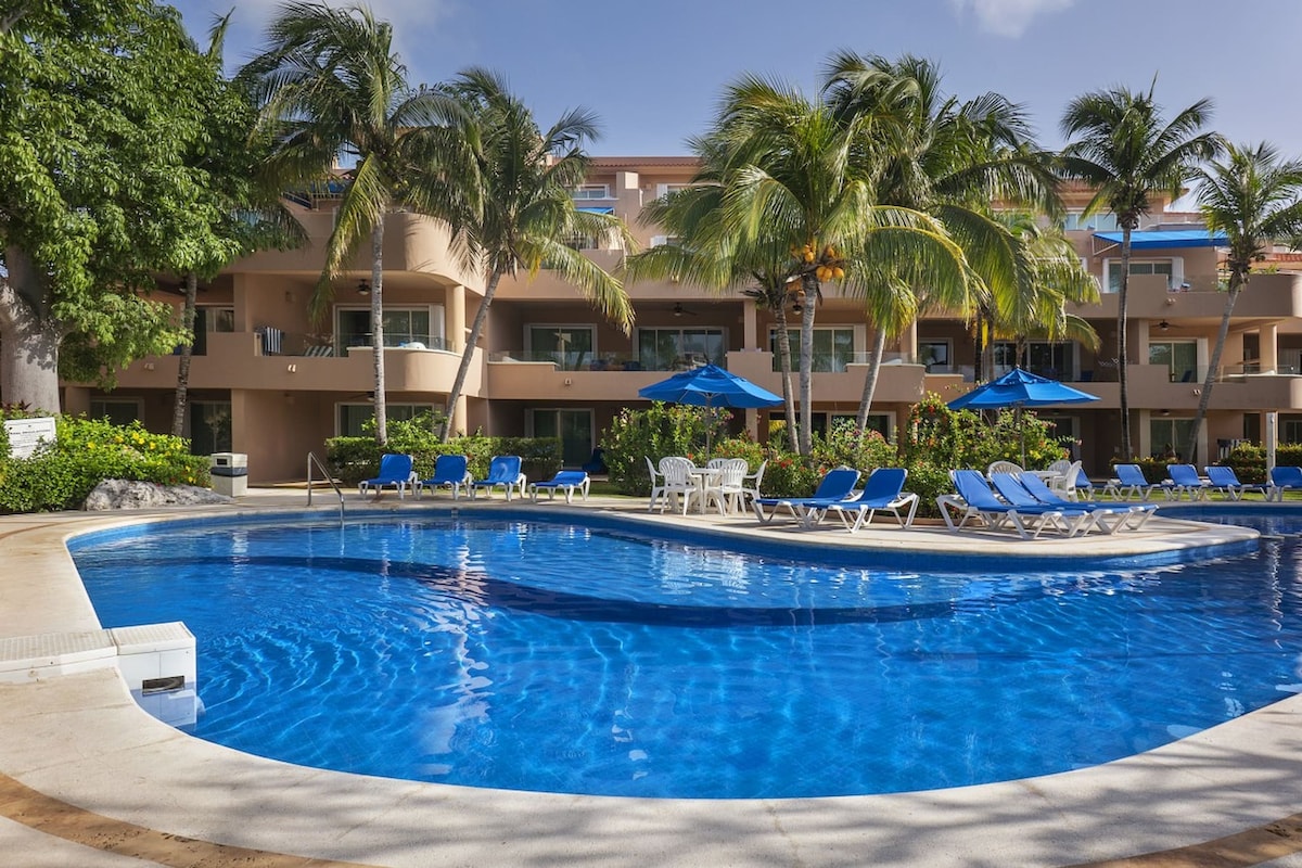 A serene swimming pool is surrounded by lush greenery and palm trees. Blue lounge chairs are arranged under large umbrellas, offering shade. The multi-level building in the background features balconies with view to the pool area, contributing to a relaxing atmosphere.