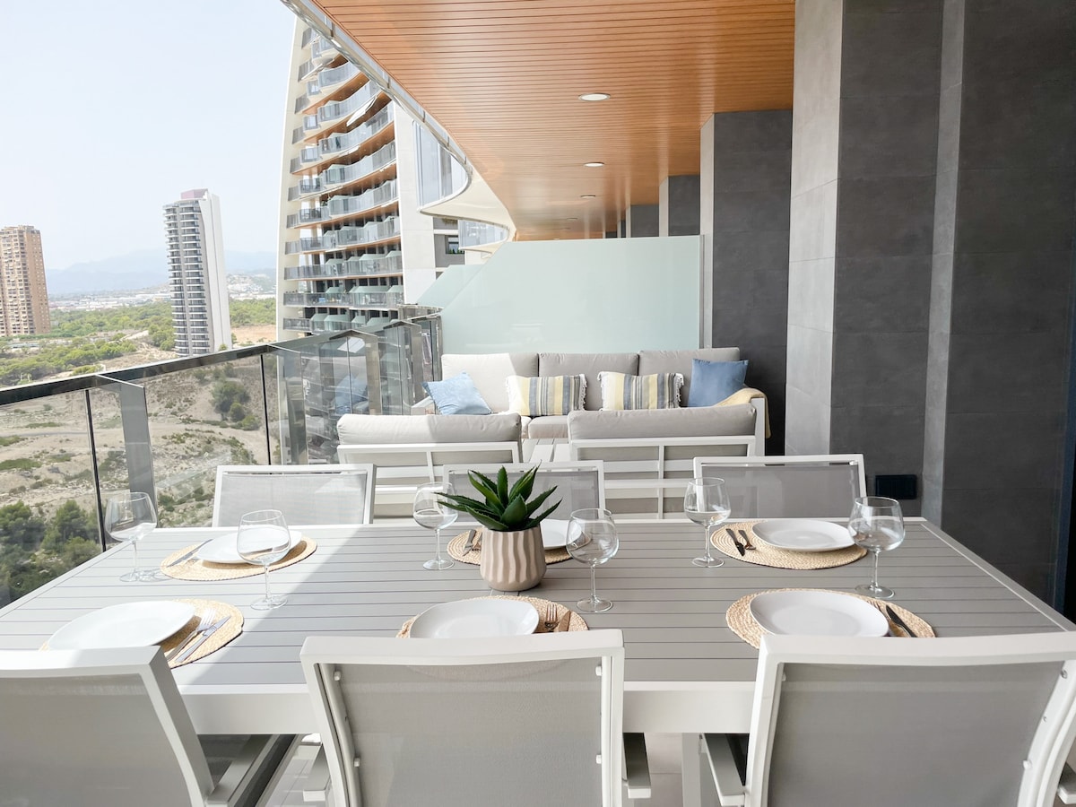 An outdoor terrace features a spacious table set for dining, complete with plates and glassware. Nearby, a comfortable seating area with soft cushions invites relaxation, complemented by a potted plant as a decorative element. The background showcases a panoramic view of the surrounding landscape.