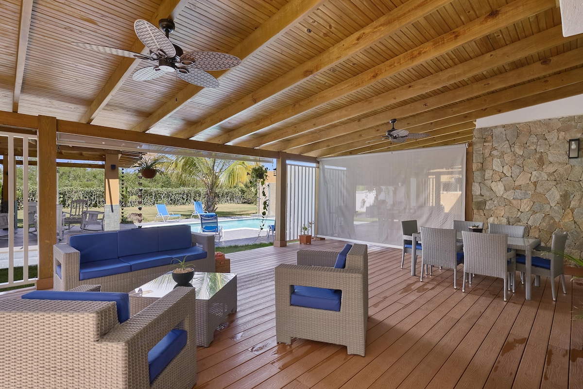 A spacious outdoor terrace features comfortable seating arrangements, including a cozy lounge area and a dining table. Ceiling fans provide a cooling breeze, while views of the pool and surrounding greenery create a relaxing setting for gatherings.