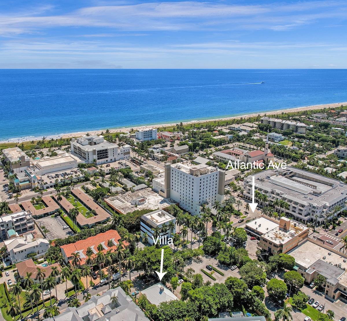 An aerial view captures the proximity of Villa Emma to the beach and the vibrant Atlantic Ave. The coastline features sandy shores meeting a blue ocean, while the lush greenery and urban landscape highlight the lively atmosphere of Delray Beach.