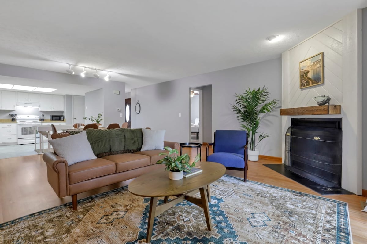 A cozy living area is featured with a brown leather sofa and a blue armchair, accompanied by a round coffee table at the center. A vibrant area rug adds warmth, while a leafy plant provides a touch of greenery. A fireplace is positioned in the corner, enhancing the welcoming ambiance.