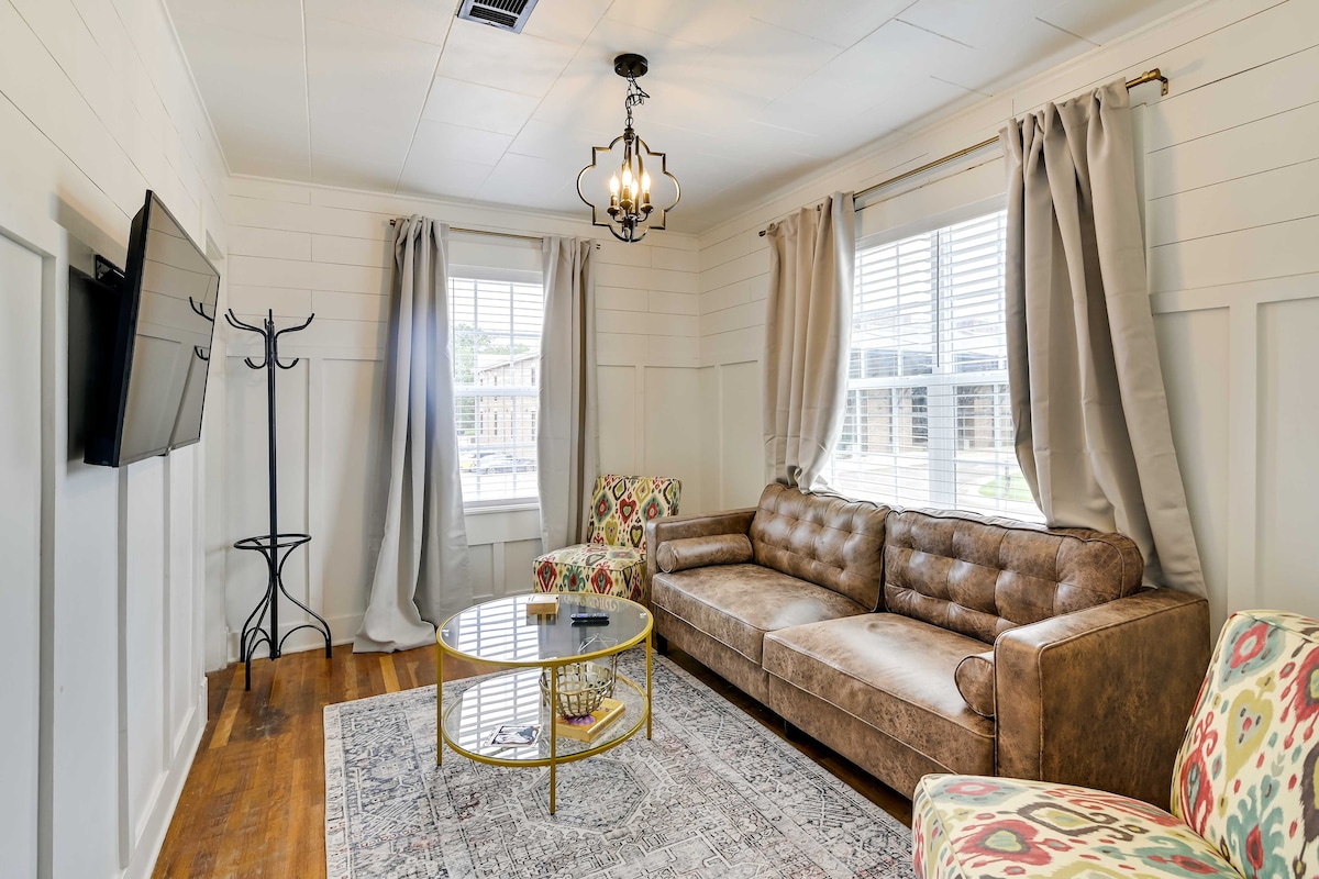 The living area showcases a sectional brown leather sofa and a patterned accent chair, inviting relaxation. A round glass coffee table sits on a soft area rug under a stylish chandelier. Natural light filters through the large windows adorned with soft curtains.