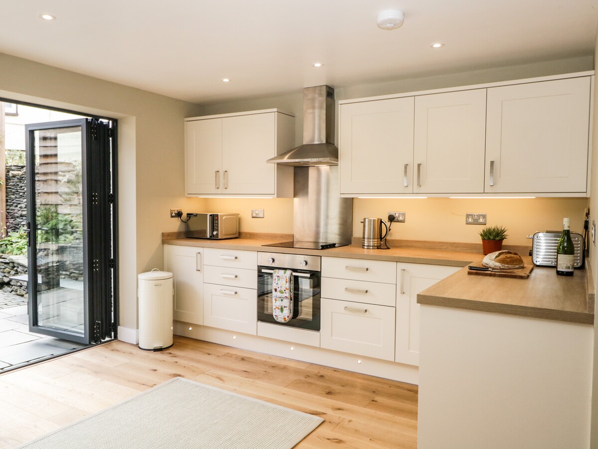 A modern kitchen is presented with sleek cabinetry and integrated appliances. An open layout connects to an outdoor space through glass doors. The countertop is complemented by under-cabinet lighting, while a light rug sits on the wooden floor.