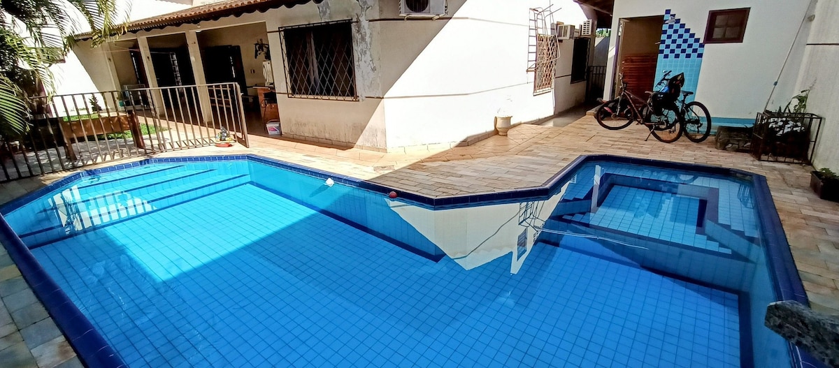 A rectangular pool features clear blue water, surrounded by tiled decking and palm greenery. The pool area is complemented by a spa section at one end. Nearby, a bicycle rests against the fence, adding to the outdoor leisure space.