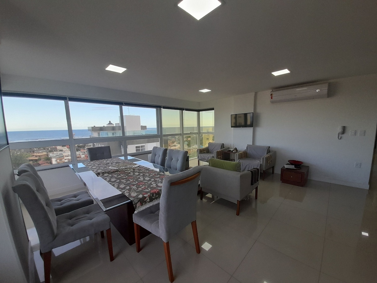 A modern living and dining area is visible, featuring a long dining table surrounded by upholstered chairs. Large windows allow natural light to fill the space, offering a view of the ocean. Comfortable seating arrangements are positioned nearby, enhancing the spacious atmosphere.