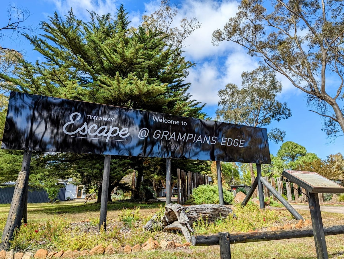 A large wooden sign welcomes guests to 'Escape @ Grampians Edge', surrounded by lush green trees and a clear blue sky. The area features natural landscaping, with a rustic wooden post design complementing the local flora.
