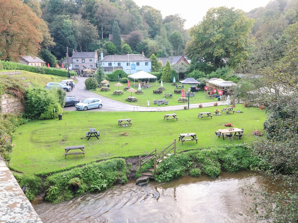 An outdoor area is presented with lush green grass, dotted with wooden picnic tables arranged neatly. Surrounding trees lend shade to the space, while a small stream flows nearby, enhancing the serene setting. Cars are parked in a designated area, contributing to the rustic scene.