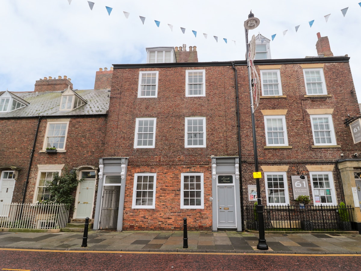 A traditional brick building is displayed, characterized by large windows and a symmetrical facade. The entrance door is centrally located, flanked by two equally spaced windows on the first and second floors. Bunting is seen strung across the top, adding a festive touch to the exterior.