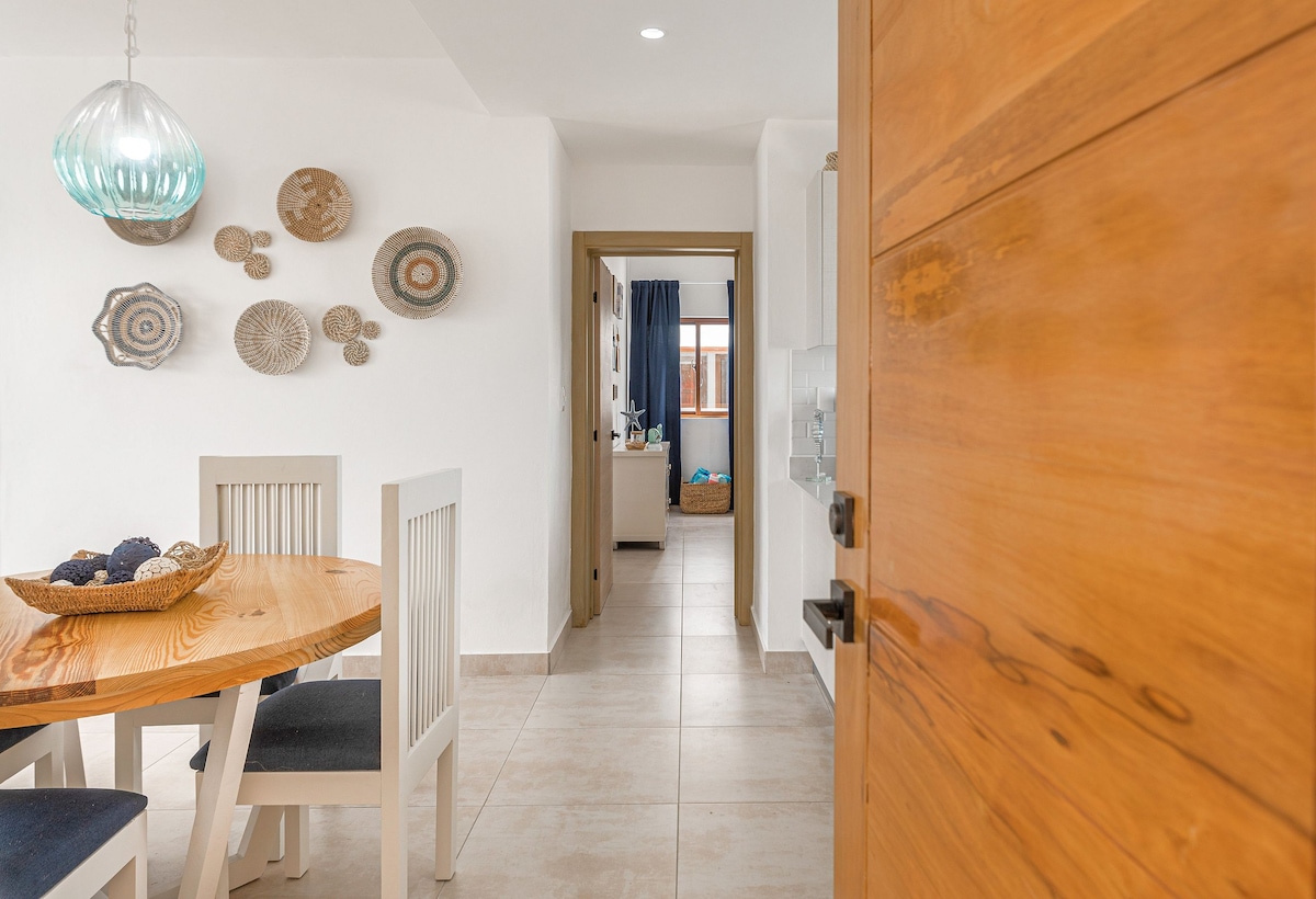 A welcoming entryway features a wooden door leading to a dining area with a round table and four white chairs. Decorative plates adorn the wall, adding charm to the light-colored space. Natural light brightens the corridor, visible in the background.