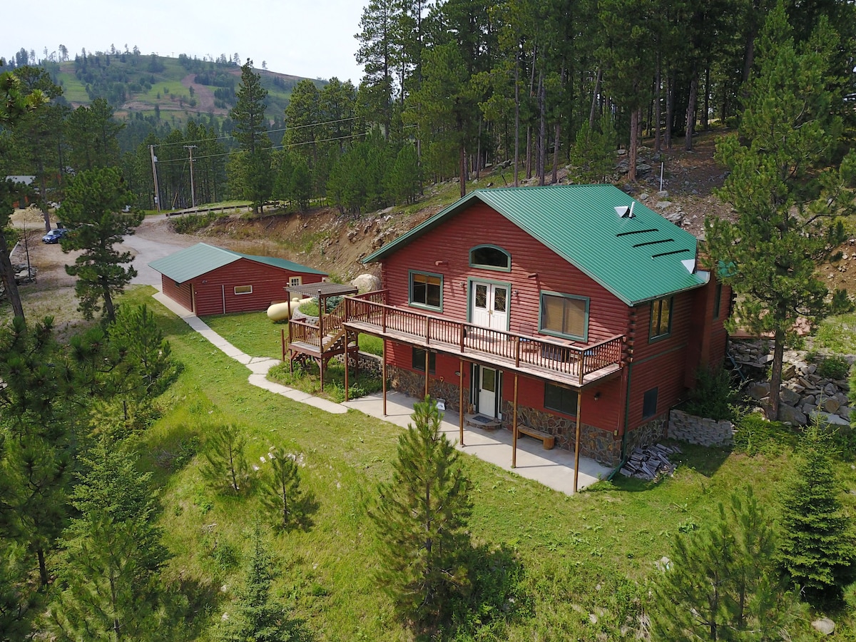 A rustic cabin is surrounded by trees and greenery, featuring a welcoming porch with a railing. The exterior is finished in warm tones, complemented by a green metal roof. A gravel road leads alongside the property, providing access to the tranquil setting.