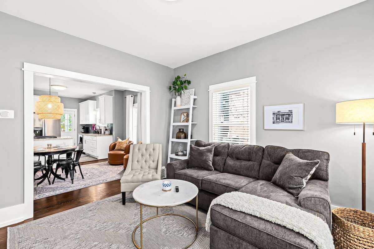 A modern living room showcases a comfortable gray sectional sofa adorned with pillows and a soft throw. A circular coffee table sits in front, accompanied by a stylish armchair. Light filters in through the windows, enhancing the inviting ambiance of the space.