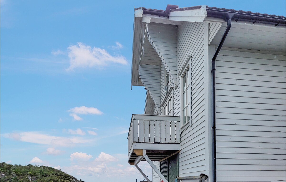 The exterior of a modern vacation home is visible, featuring clean white lines and a sloped roof. A balcony extends out from the upper level, offering views of the surrounding landscape. The backdrop includes a clear blue sky with soft clouds.