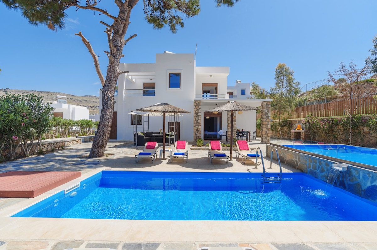 A contemporary villa features a private pool surrounded by sun loungers and parasols. The spacious terrace is highlighted by a flowering garden and shaded areas. The villa's exterior is a blend of white walls and natural stone, with large windows providing views of the pool area.