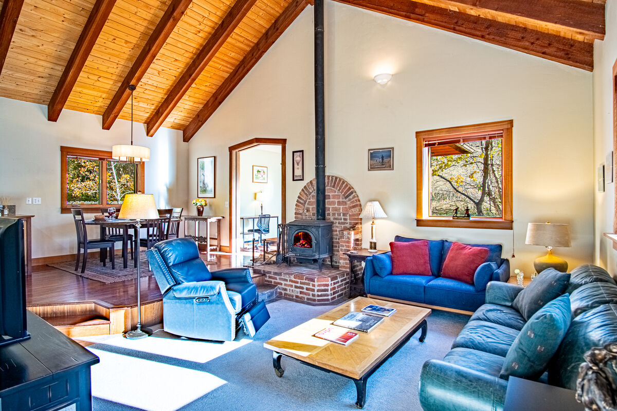 A spacious living area features vaulted wooden ceilings and large windows that allow natural light to brighten the room. A wood-burning stove is set within a brick surround, and comfortable seating options include a dark blue sofa and a light blue chair. A wooden dining area is visible in the background.