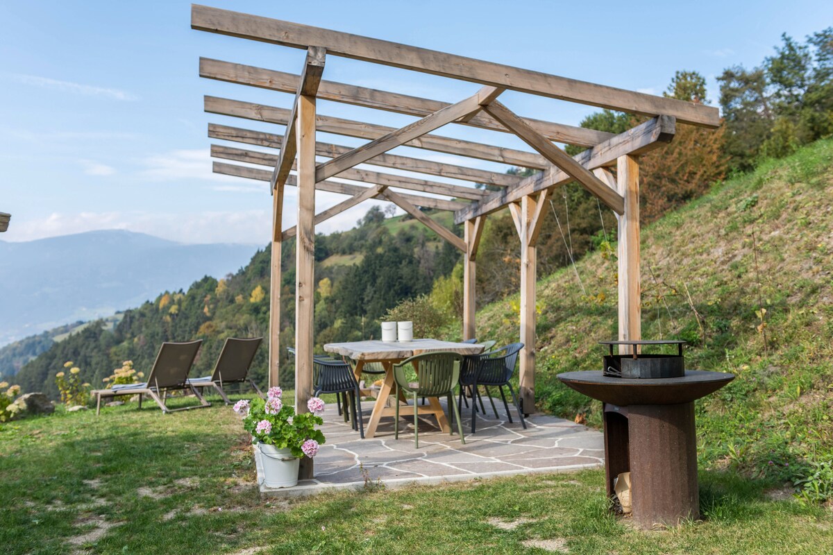 An outdoor seating area is presented under a wooden pergola, featuring a large table surrounded by chairs. A charcoal barbecue is visible nearby, along with a flower pot containing pink blossoms. The backdrop showcases rolling hills and mountains under a clear sky.